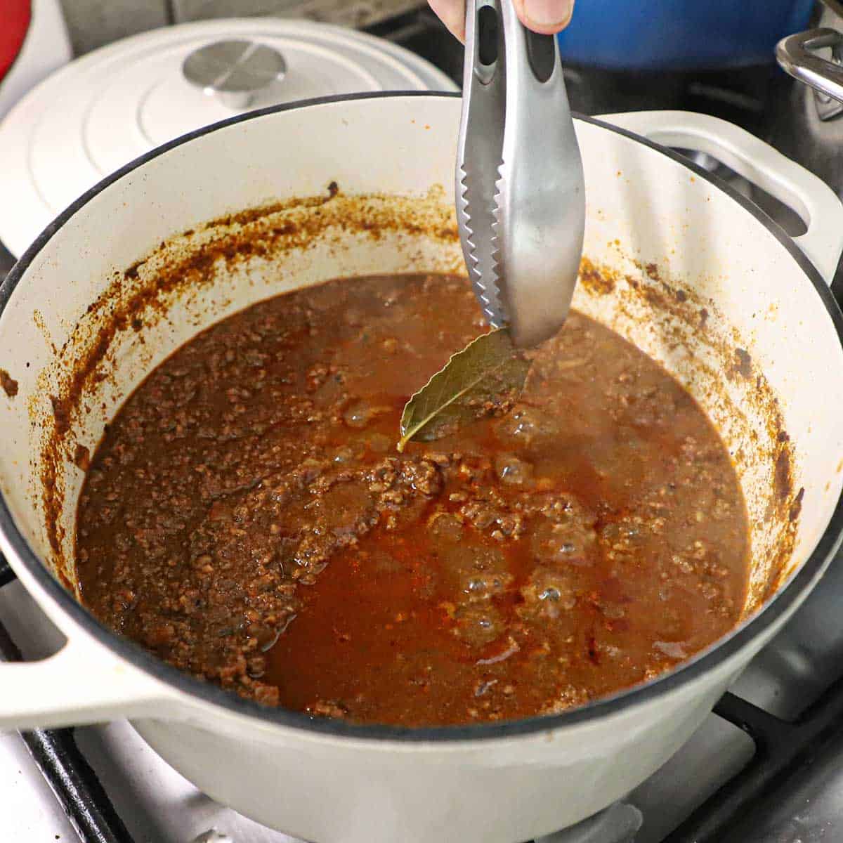 A person using a pair of metal tongs to remove a bay leave from a pot of simmering Cincinnati chili.