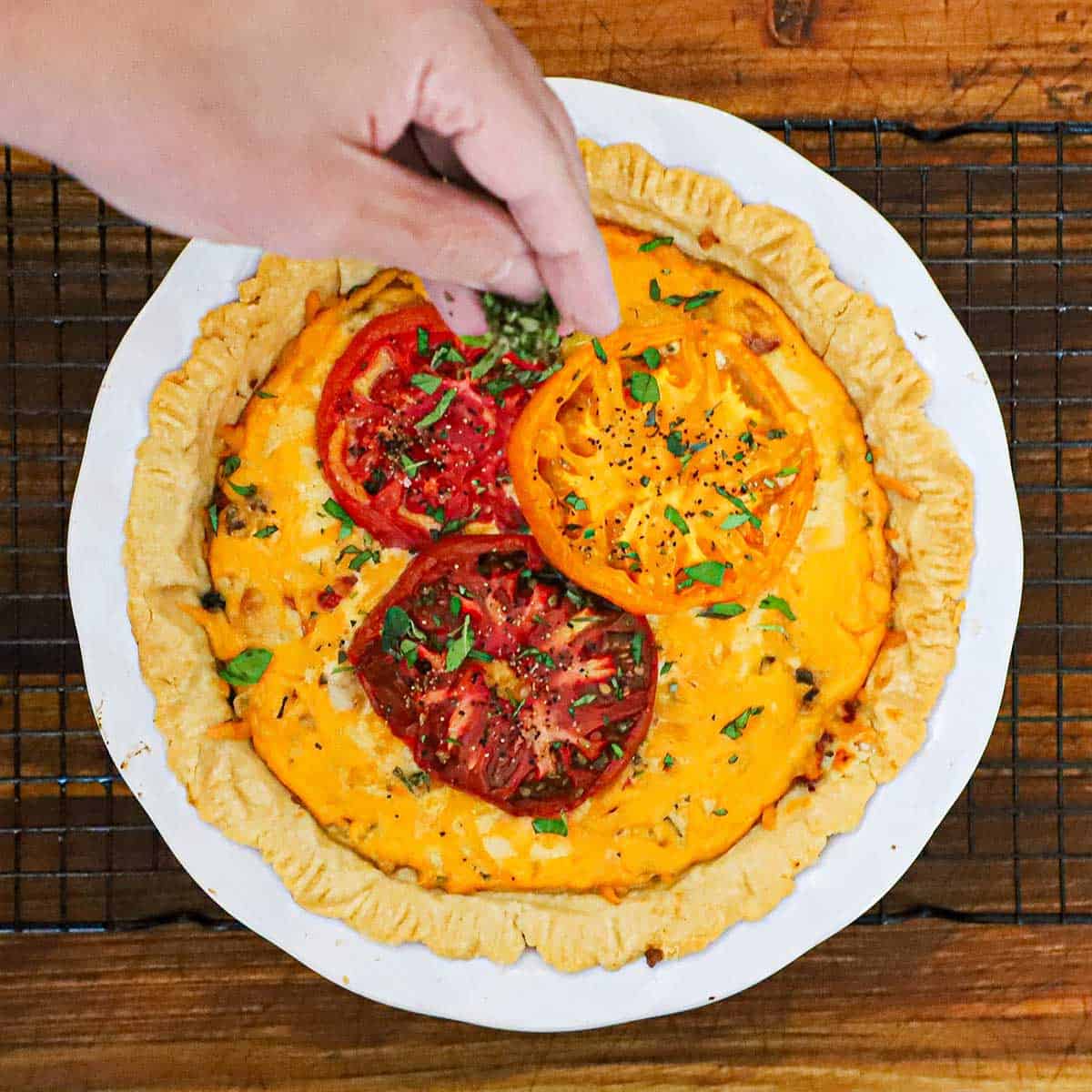 A person sprinkling chopped fresh herbs over a baked Southern tomato pie resting on a baking rack on a wooden cutting board.