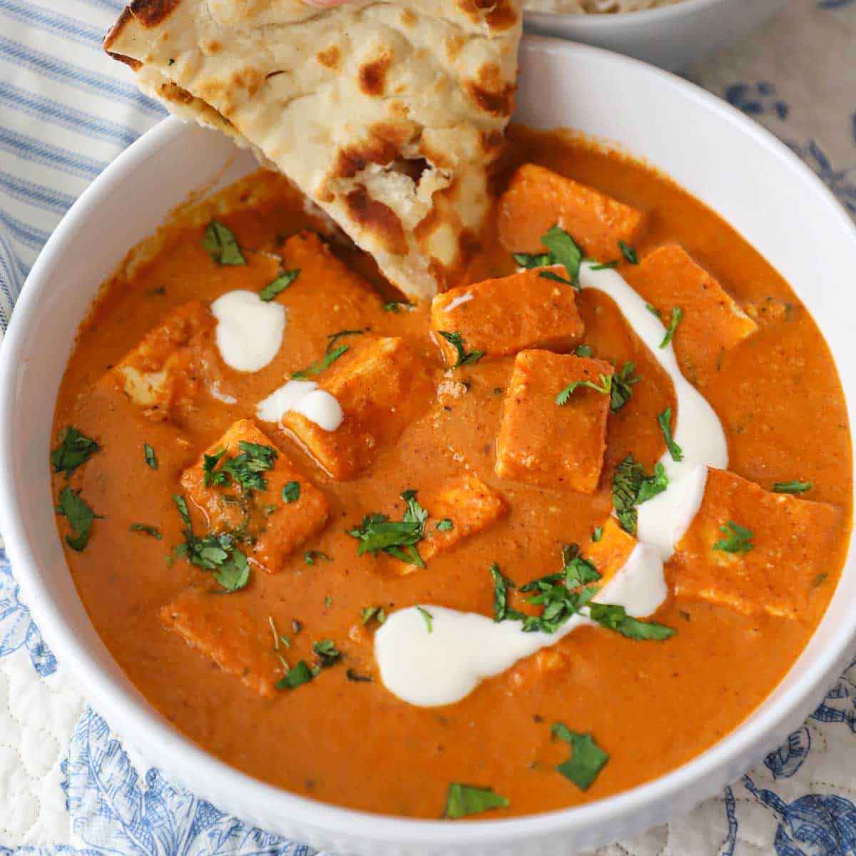 A white serving bowl filled with a serving of paneer butter masala topped with a streak of cream, chopped cilantro, and a triangle of naan in the side of the masala.