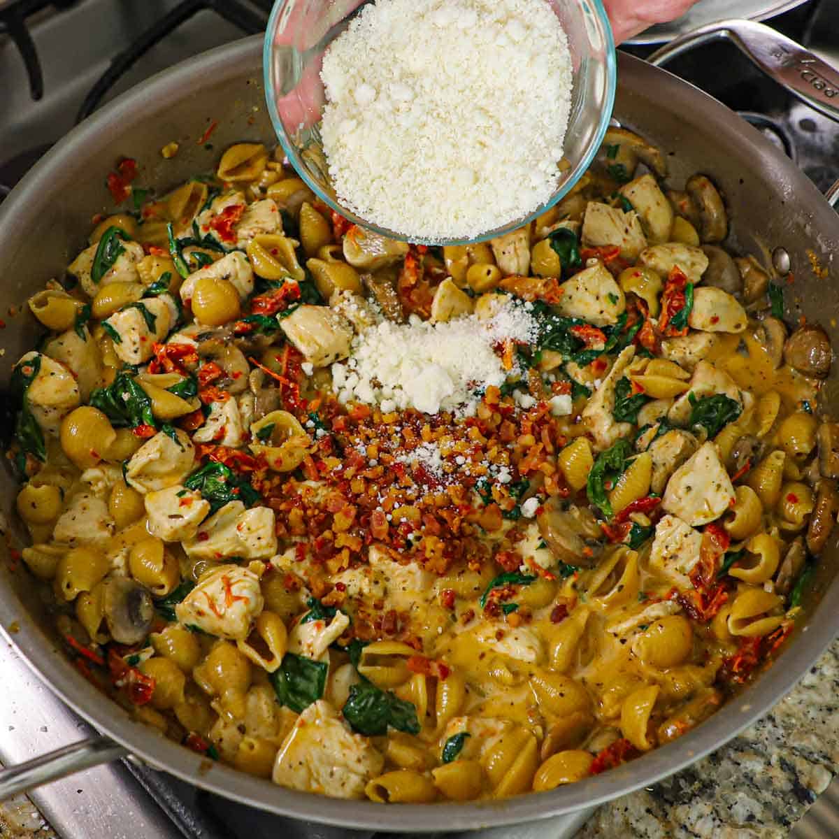 A person dumping grated Parmesan cheese from a small glass bowl into a pot filled with a simmering Tuscan sauce with cooked pasta, cubed chicken, crispy pancetta, and wilted spinach.