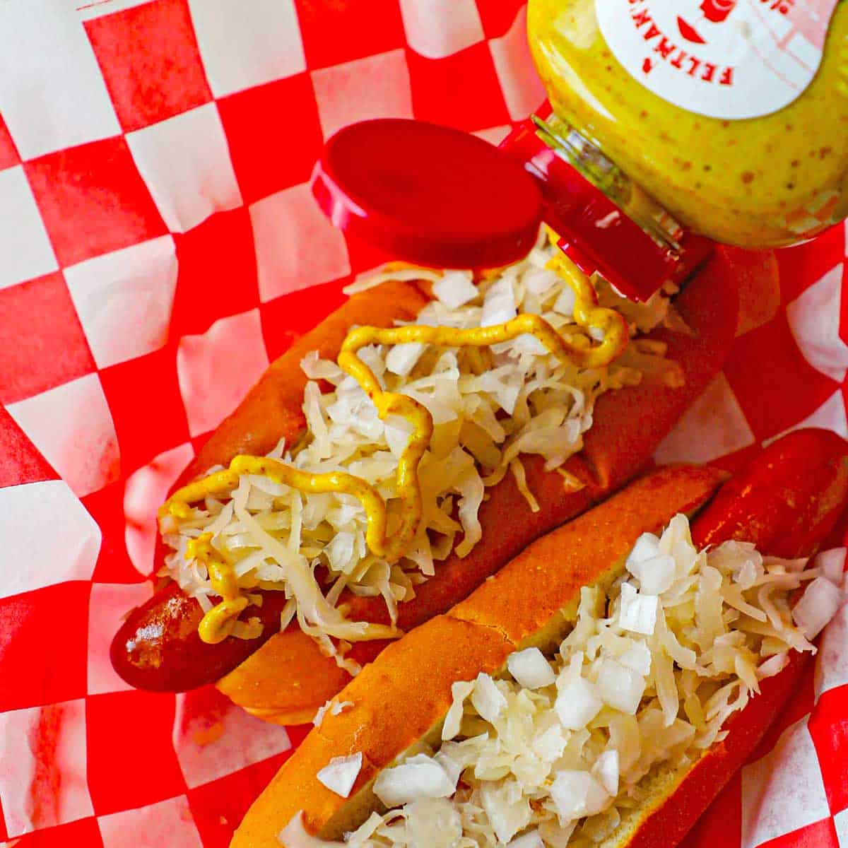 A person adding a zig zag line of deli style mustard from a bottle onto two Coney Island hot dogs that are topped with sauerkraut and chopped onions.