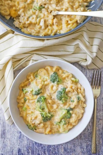 A white bowl filled with a serving of creamy mac and cheese with chicken and broccoli sitting next to a large skillet filled with the same.