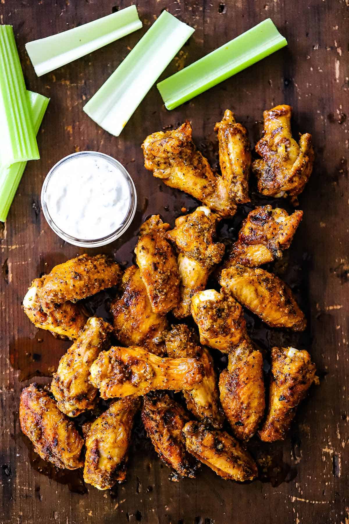 A pile of grilled chicken wings with buffalo sauce resting on a wooden board next to a jar of blue cheese dressing and cut strips of celery.