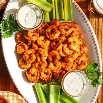 A white oval platter filled with a pile of grilled buffalo shrimp with two small jars filled with blue cheese dressing and celery sticks on each side of the dish.