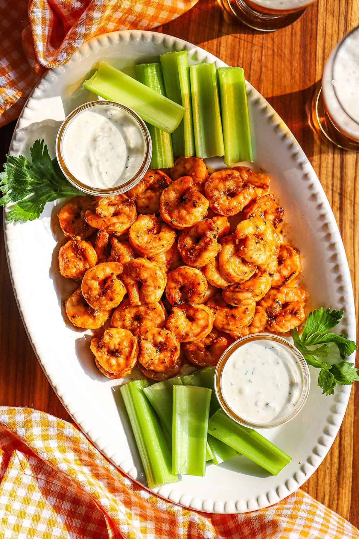A white oval platter filled with a pile of grilled buffalo shrimp with two small jars filled with blue cheese dressing and celery sticks on each side of the dish.