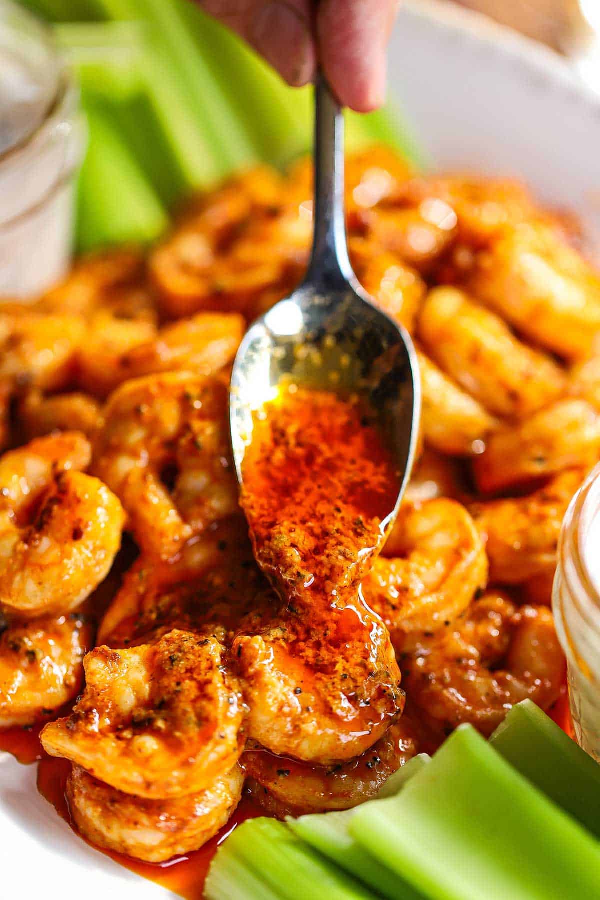 A person using a spoon to drizzle Buffalo sauce over a platter filled with grilled buffalo shrimp with celery sticks resting on the sides of the dish.