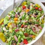 A close-up view of a large white platter filled with an Italian chopped salad with homemade vinaigrette with two serving utensils inserted into the side of the salad.