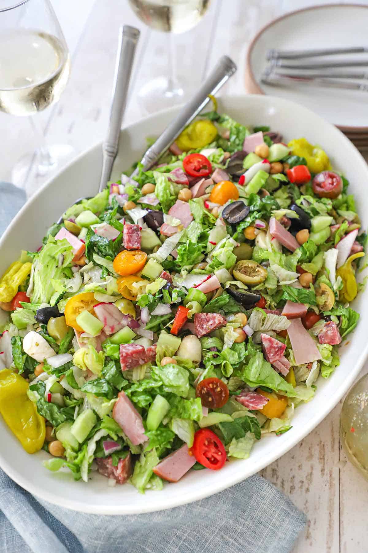 A large white platter filled with and Italian chopped salad with homemade vinaigrette with two serving utensils inserted into the side of the salad.