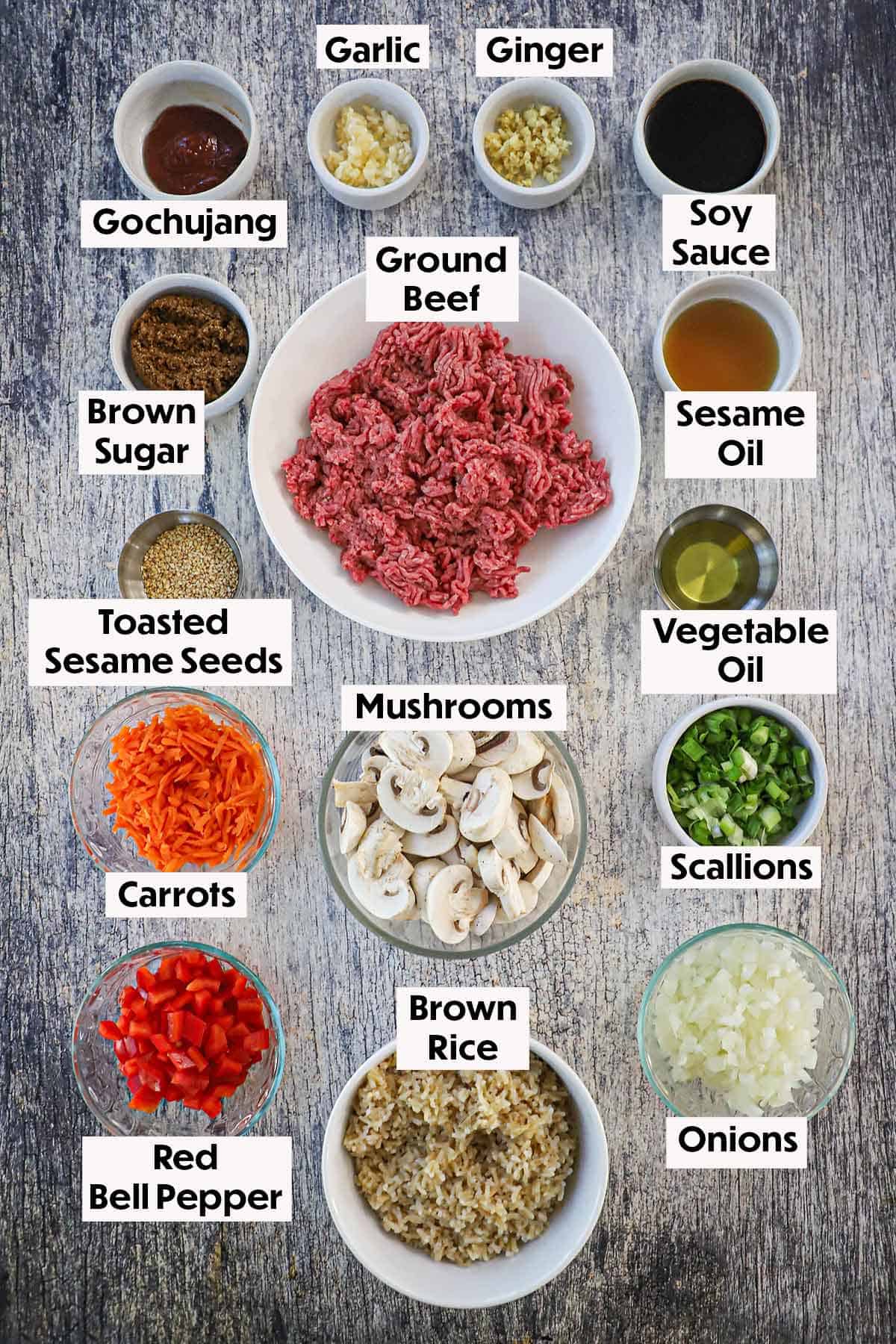 An arrangement of ingredients for Korean ground beef stir-fry on a wooden grey background including ground beef, mushrooms, carrots, bell peppers, onions, scallions, rice, garlic, gingers, and seasonings.
