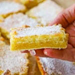 A person holding a square piece of a Gooey Butter Cake that has been dusted with powdered sugar and is near other cut squares of the cake.