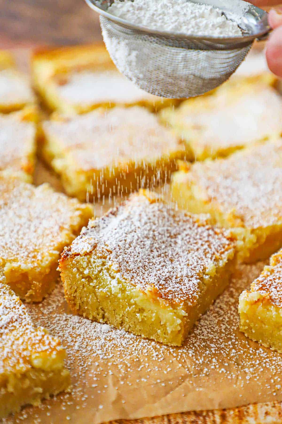 A person using a small sifter to dust powdered sugar over squares of Gooey Butter Cake resting on brown paper.