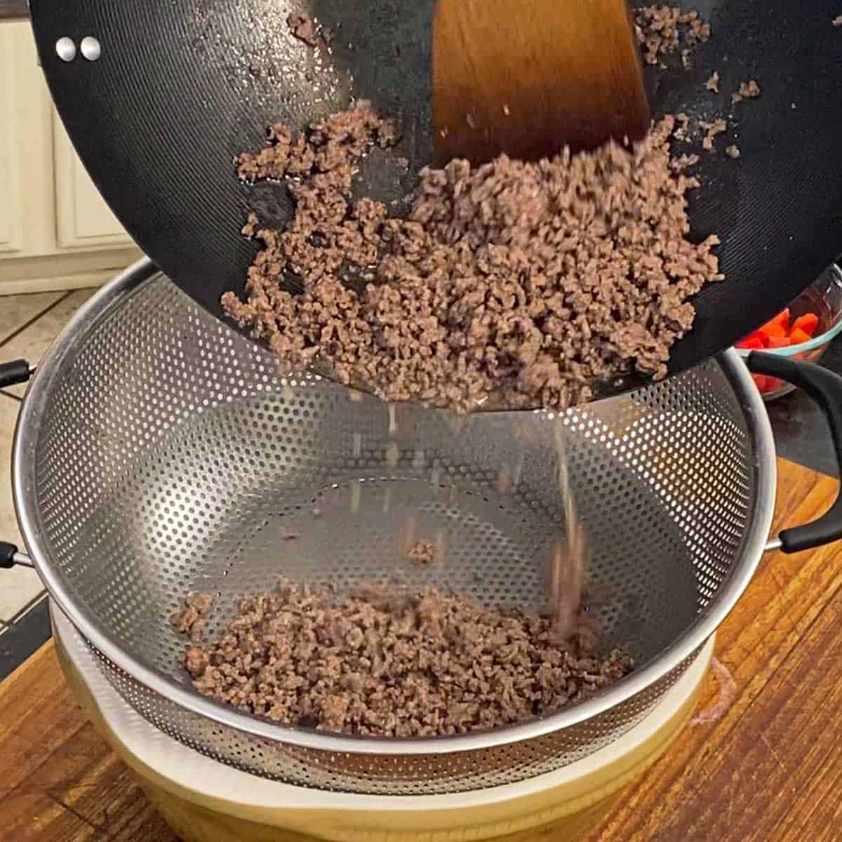 A person transferring cooked ground beef into a large colander resting on top of a ceramic bowl that is catching the rendered grease from the meat.