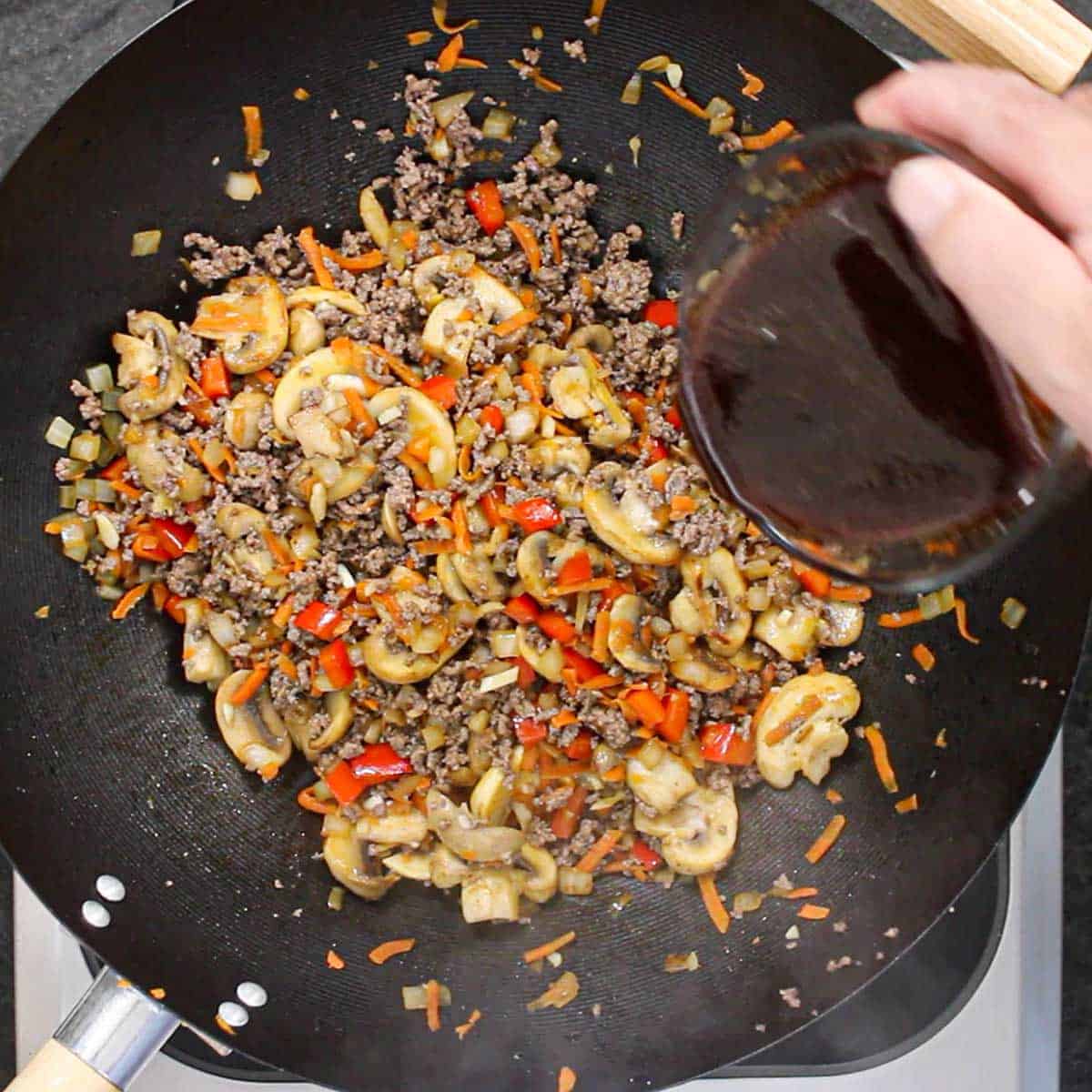 A person pouring a Korean stir-fry sauce from a small glass bowl into a large wok that is filled with simmering cooked ground beef, sautéed mushrooms, and soft chopped bell peppers and shredded carrots.