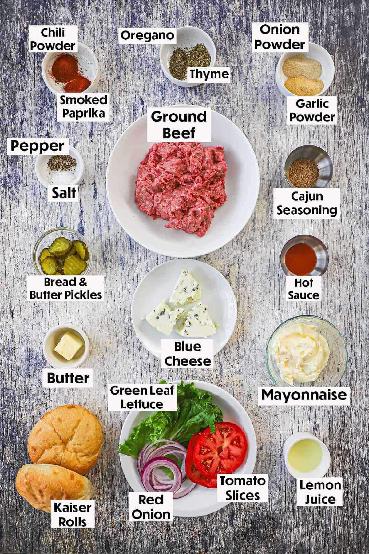 An arrangement of ingredients for a black and bleu burger on a wooden grey background including ground beef, blue cheese, blackening seasonings, pickles, buns, mayonnaise, lemon juice, lettuce, red onion, and tomato slices.