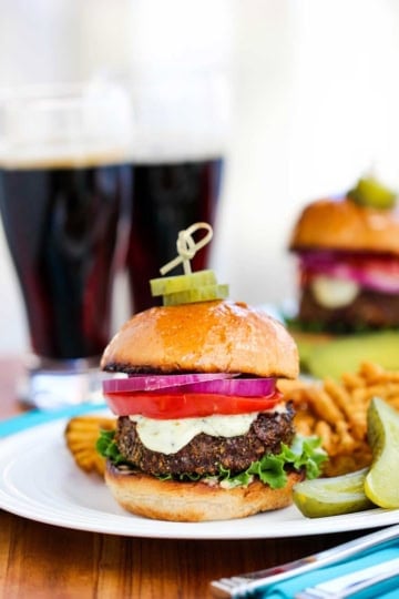 A black and bleu burger with two pickle slices stuck in the top of the bun with a tooth pick all on a white plate next to crosshatch fried and two spears of pickles and two glasses of dark beer in the background.