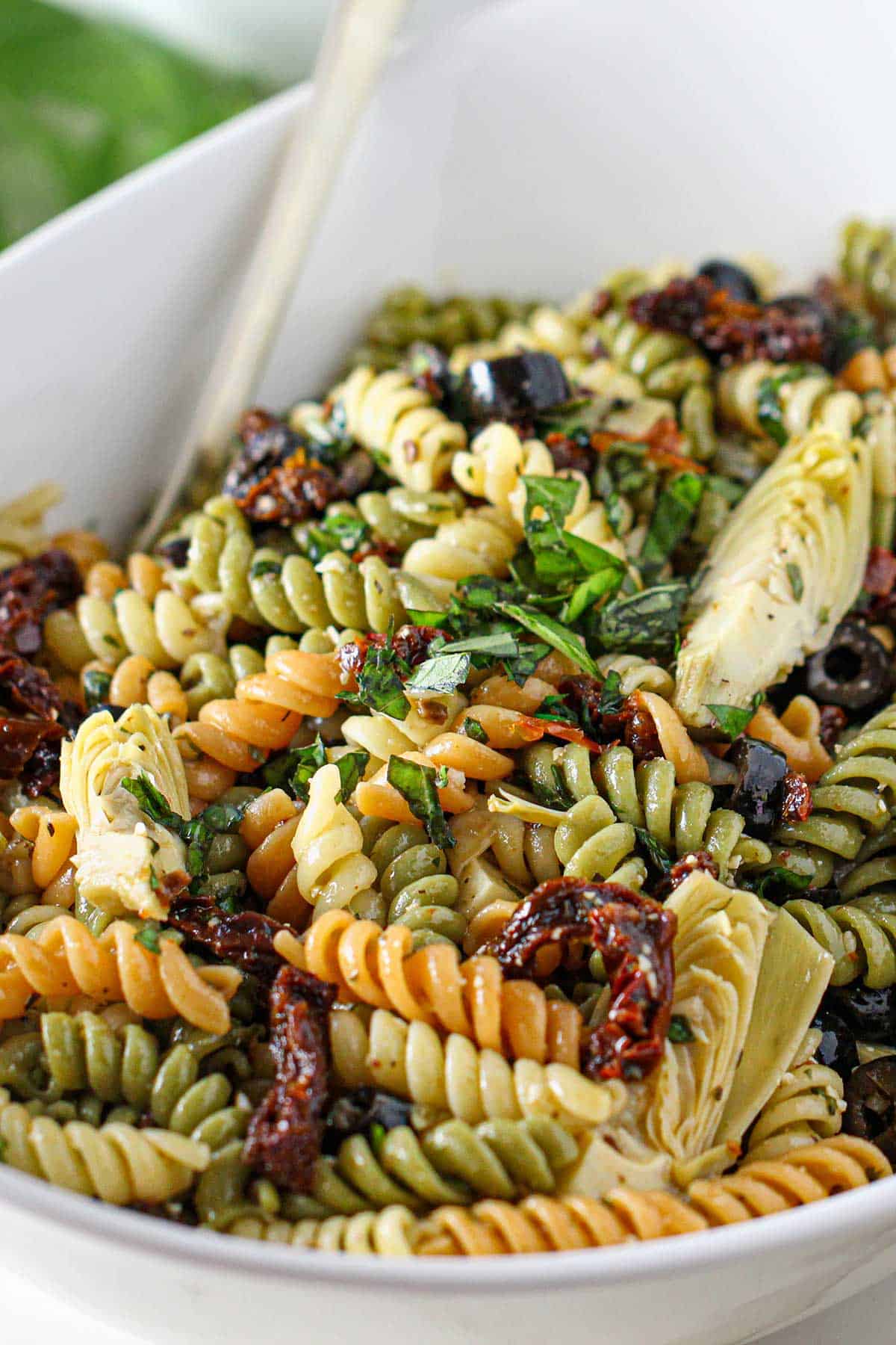 A close-up view of a classic pasta salad in a white oval serving bowl topped with chopped basil.