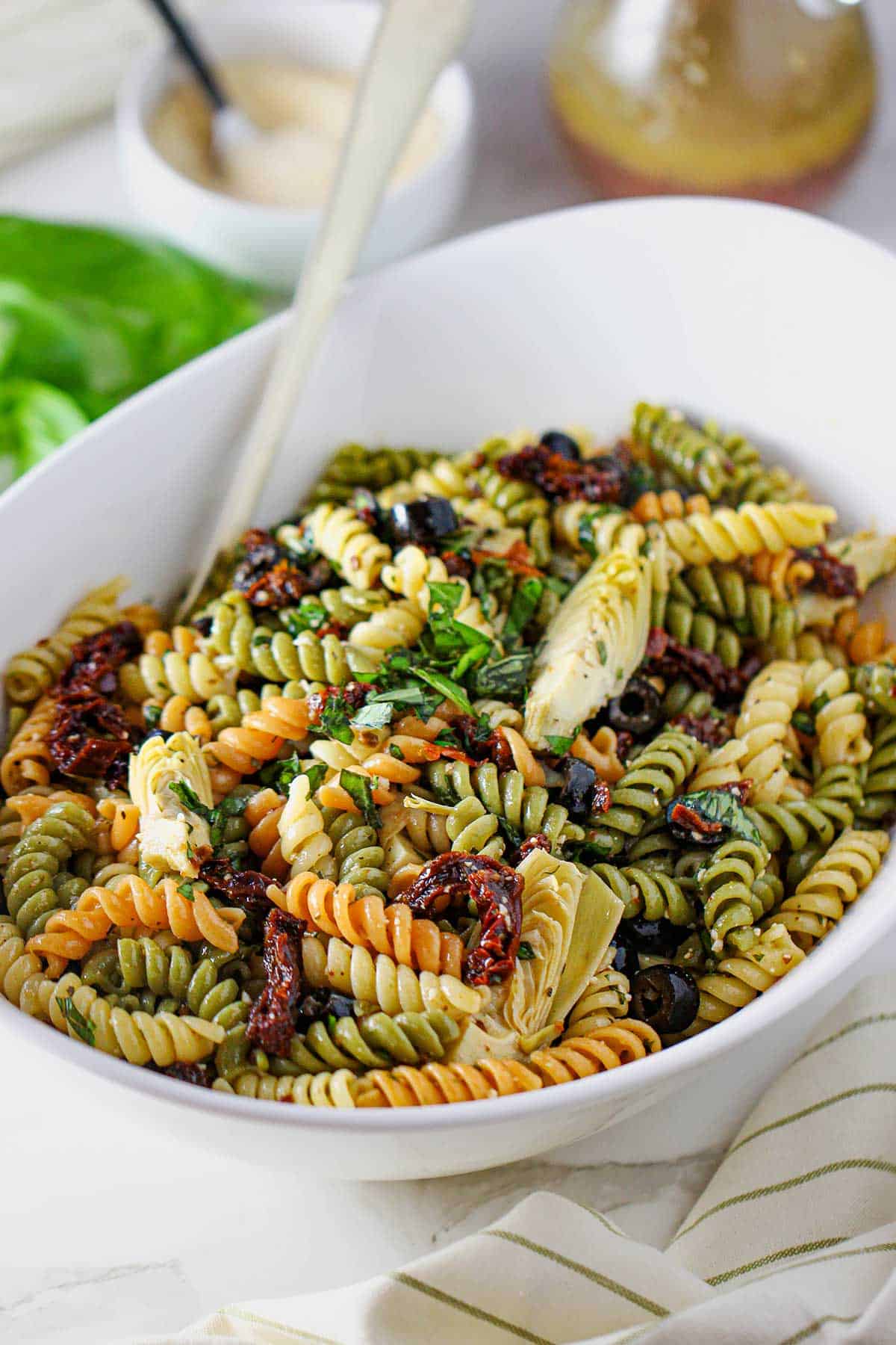 A white oval serving bowl filled with classic pasta salad including artichoke hearts, sun-dried tomatoes, black olives, tricolor rotini pasta, basil, tossed in homemade Italian dressing.