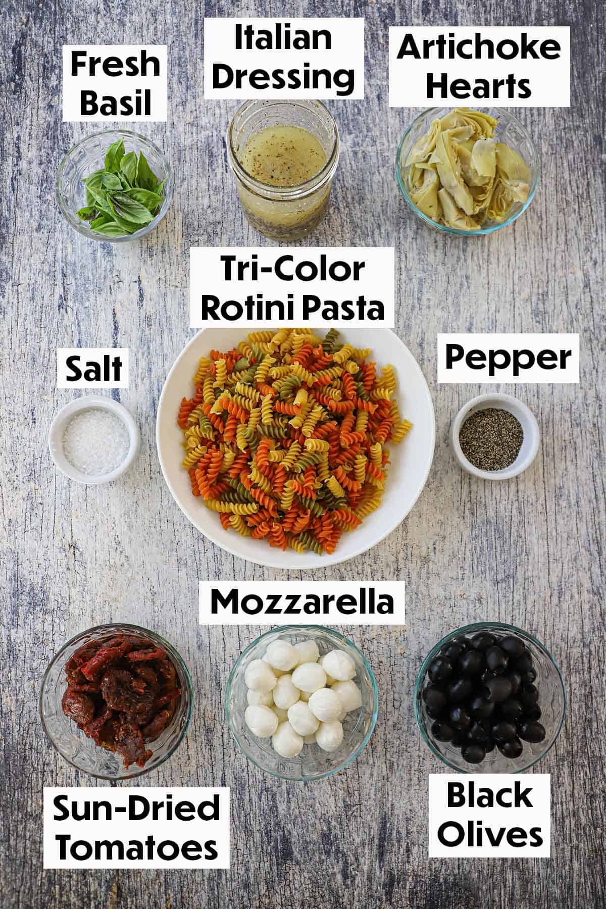 An arrangement of ingredient for Classic Pasta Salad on a grey wooden background including tricolor rotini pasta, basil, artichoke hearts, sun-dried tomatoes, mozzarella balls, olives, salt, pepper, and Italian dressing.