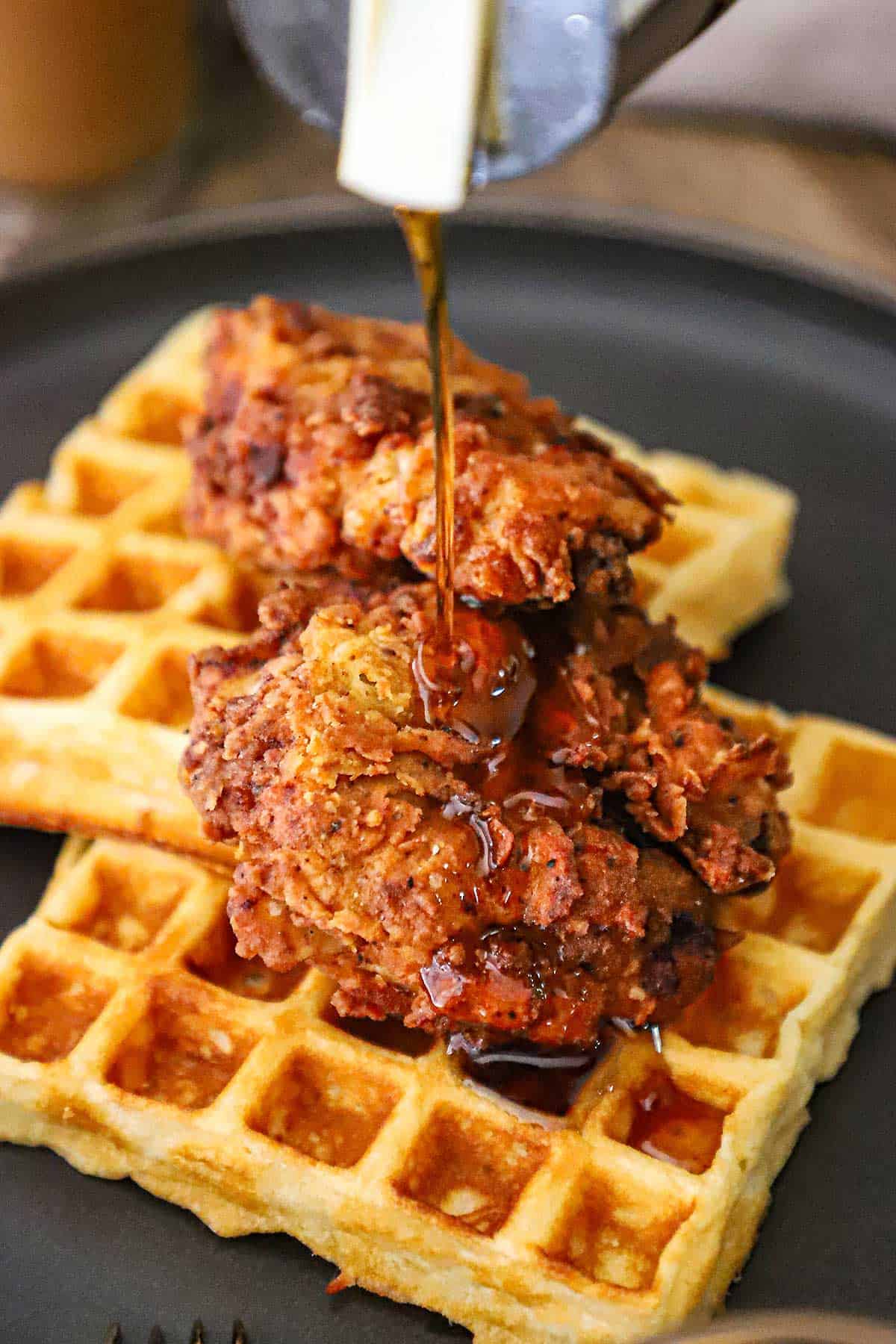 A person pouring maple syrup from a syrup bottle over a serving of crispy chicken and waffles on a black dinner plate.