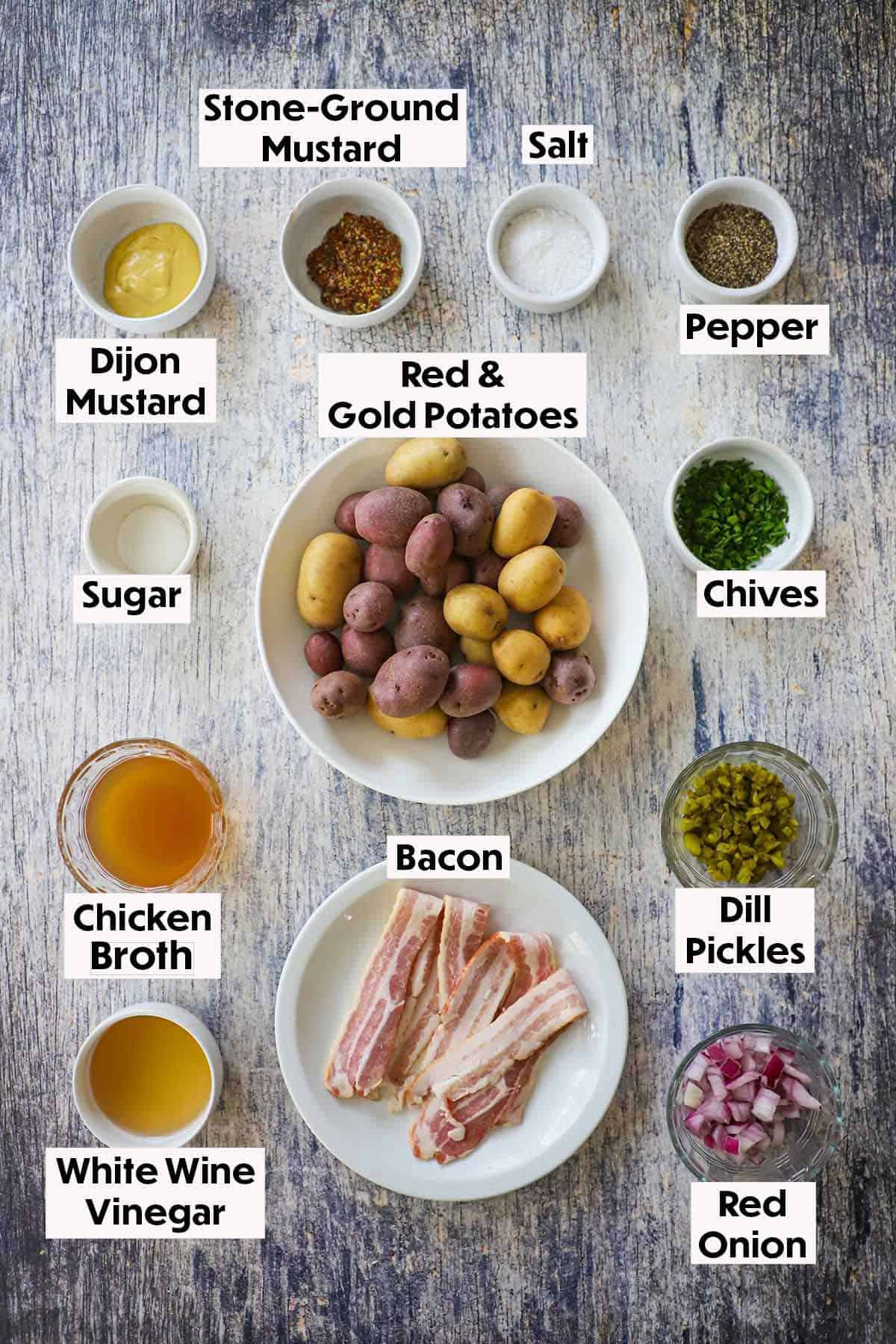 An arrangement of ingredients for best German potato salad on a grey wooden background including red and gold potatoes, Dijon mustard, stone-ground mustard, sugar, chives, chicken broth, vinegar, bacon, pickles, and onions. 