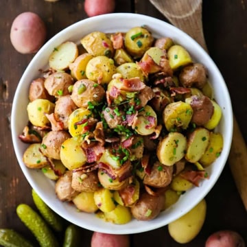 An overhead view of a white serving bowl filled with a mound of Best German Potato Salad that is topped with chopped crispy bacon and snipped chives.