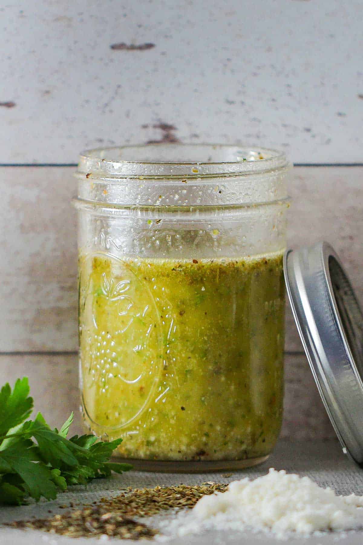 A Mason jar filled with homemade Italian dressing with a small pile of grated parmesan cheese, dried herbs, and fresh parsley around the jar.
