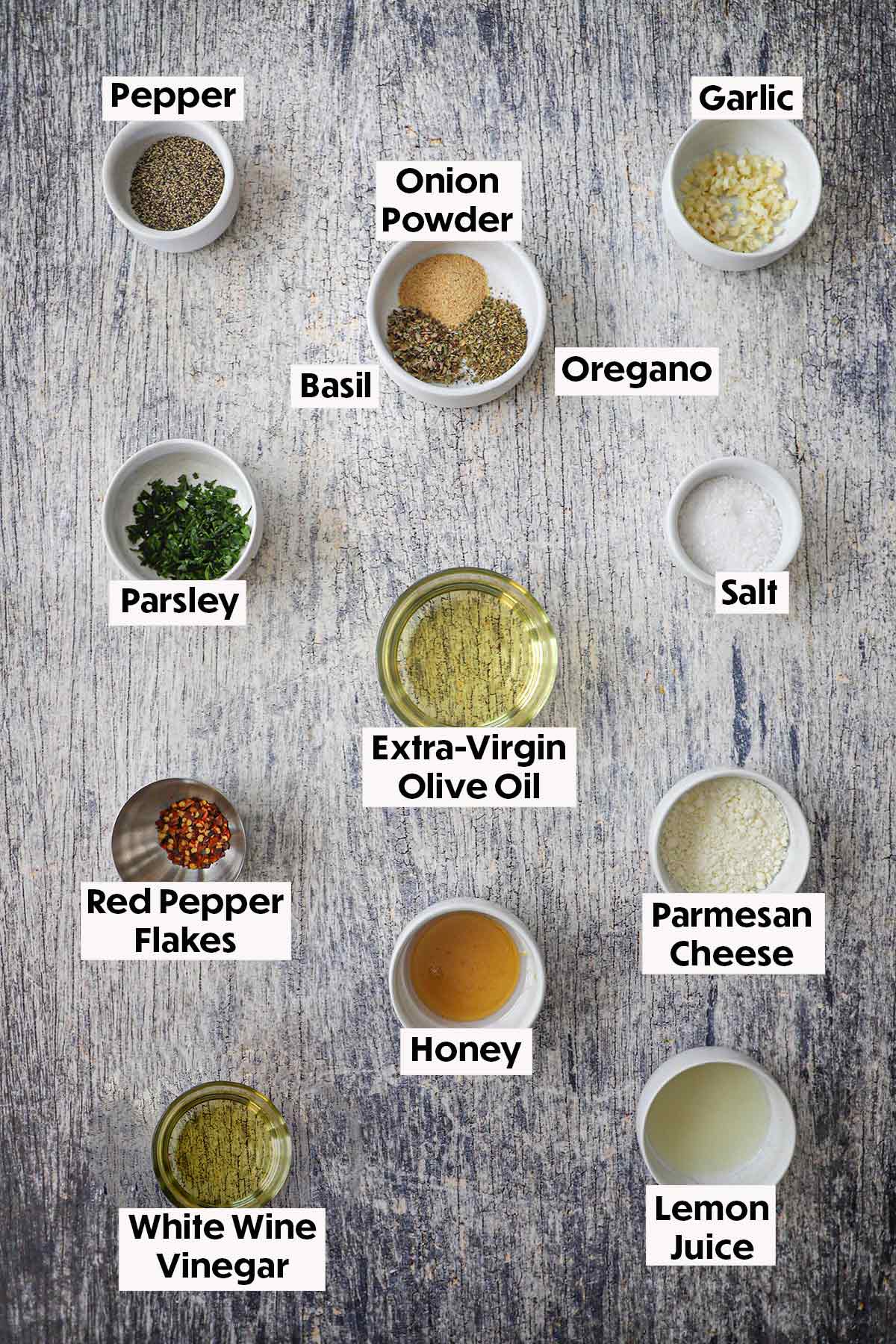 An arrangement of ingredients on a grey wooden background for homemade Italian dressing including olive oil, garlic, parsley, honey, Parmesan cheese, honey, vinegar, red pepper flakes, and seasonings.