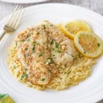 A plate filled with a serving of pan-fried cod with lemon butter sauce resting on cooked orzo pasta and two lemon wedges off to the side of the dish.