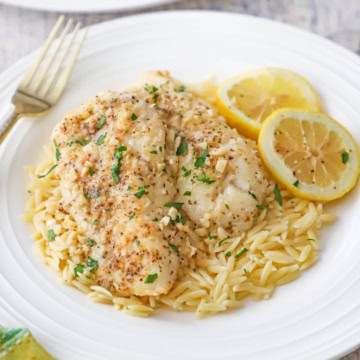 A plate filled with a serving of pan-fried cod with lemon butter sauce resting on cooked orzo pasta and two lemon wedges off to the side of the dish.