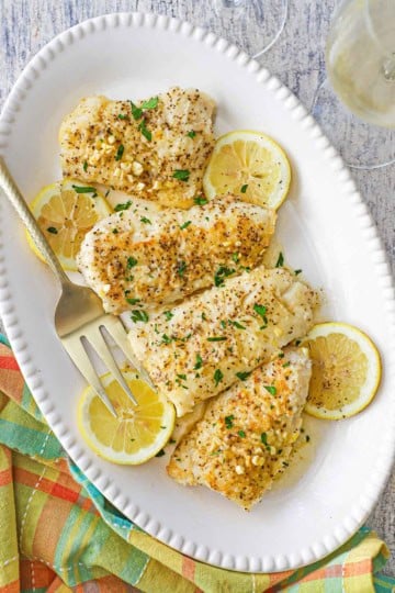 An overhead view of four pan-fried filets of cod topped with a lemon butter sauce all on a beaded oval platter with lemon slices scattered around the fish.