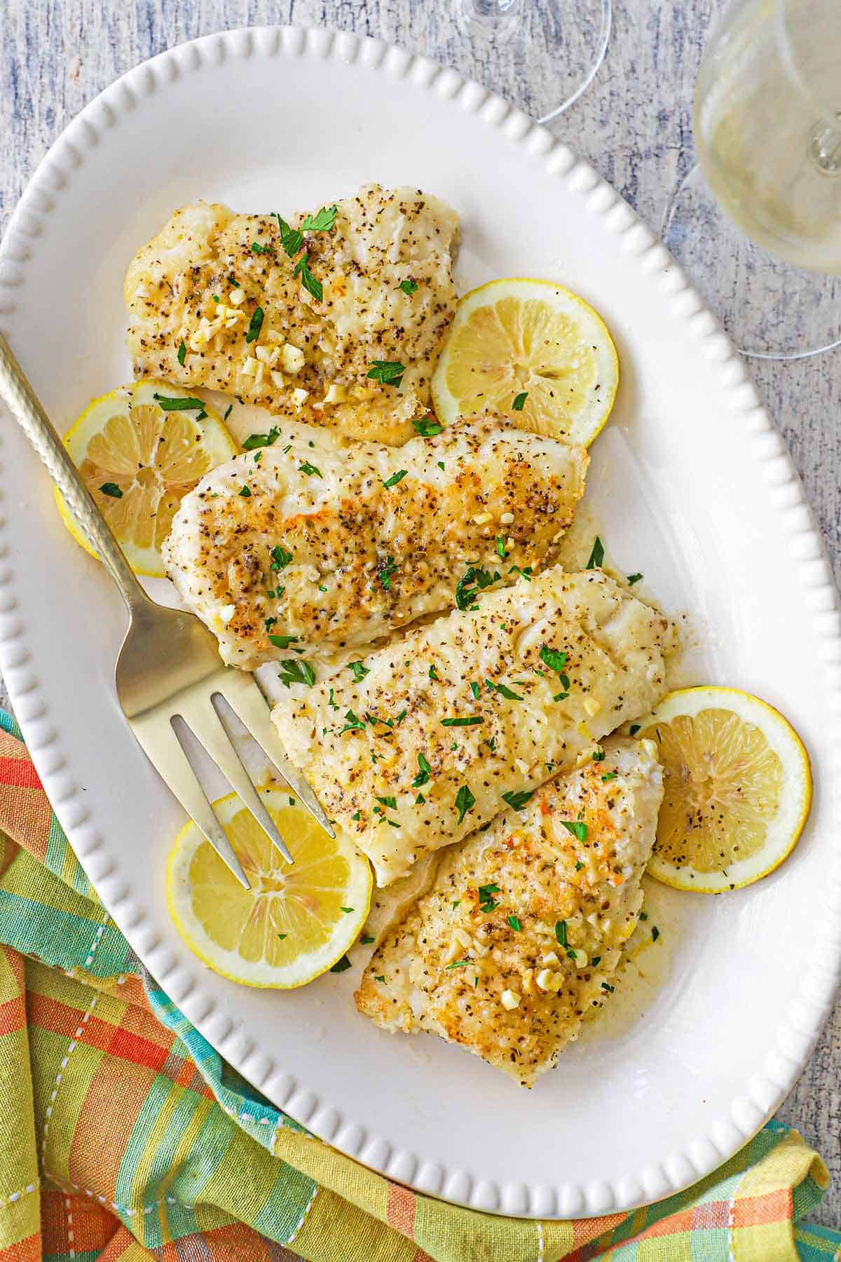 An overhead view of four pan-fried filets of cod topped with a lemon butter sauce all on a beaded oval platter with lemon slices scattered around the fish.