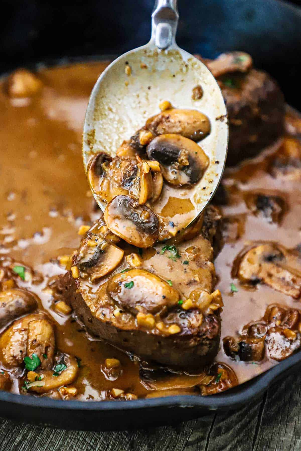 A serving spoon pouring a mushroom brown gravy sauce of a fully cooked filet mignon in a cast-iron skillet that is filled with classic steak Diane.