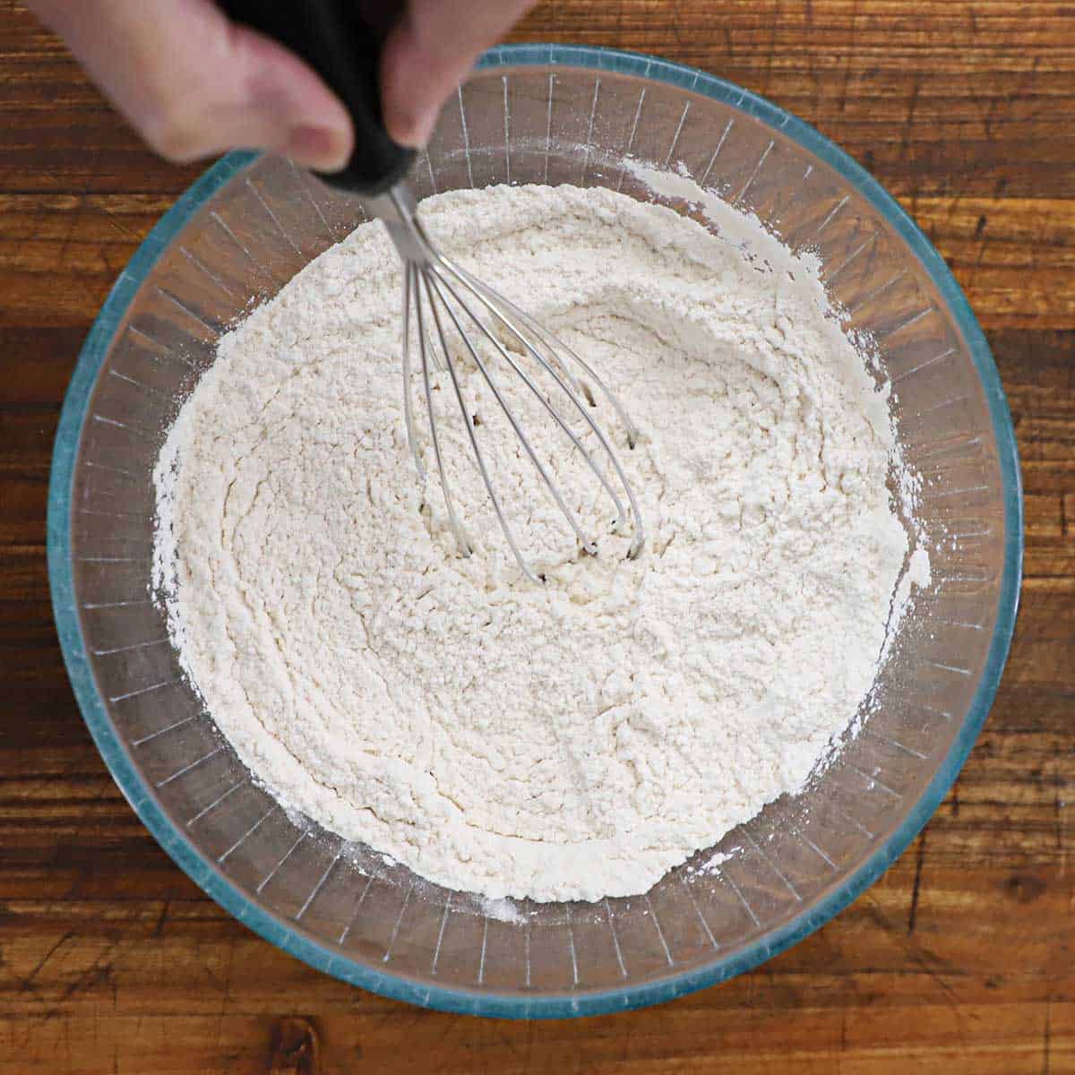 A person using a whisk to mix flour, salt, and baking powder in a glass bowl on a wooden cutting board.
