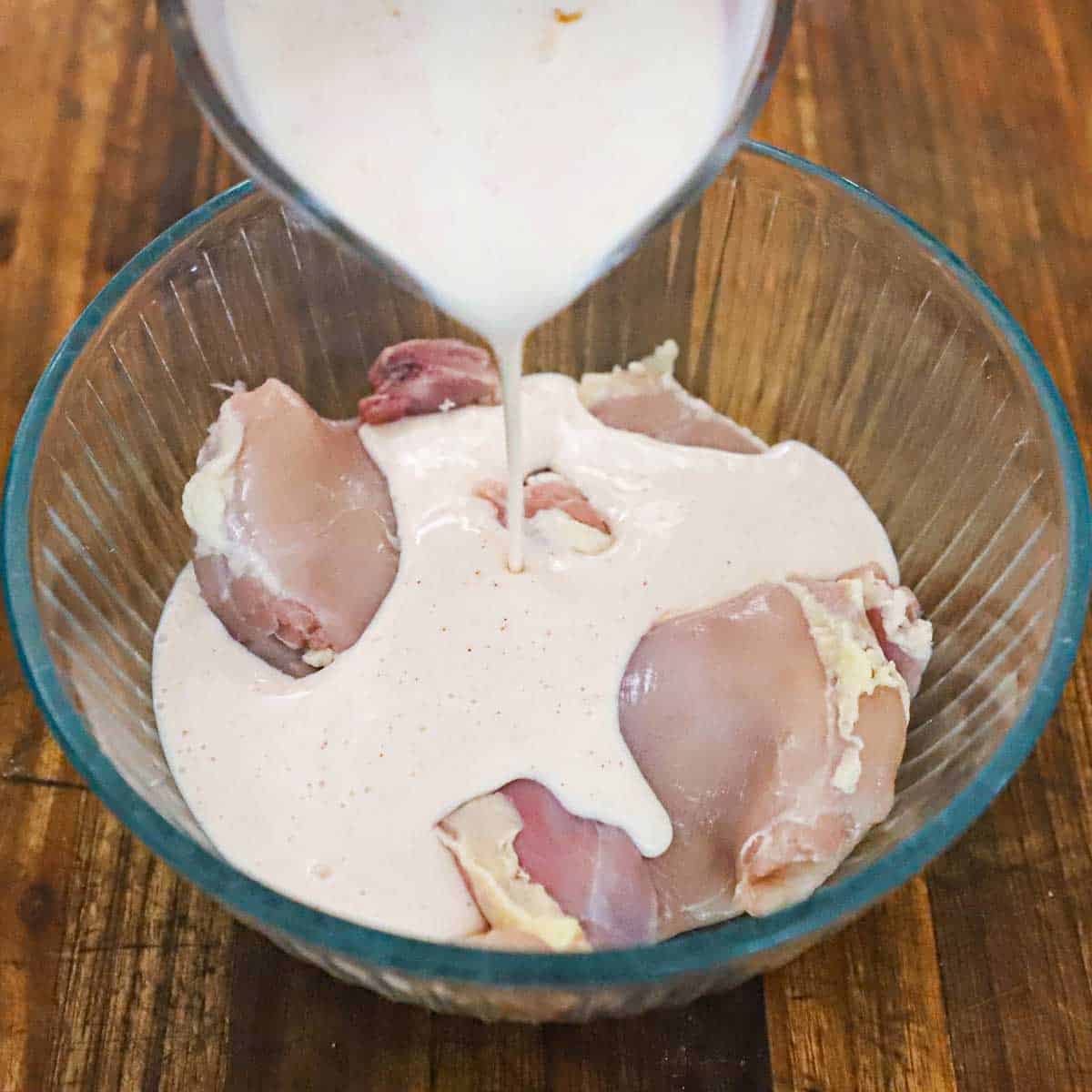 A person pouring seasoned buttermilk from a glass measuring cup into a glass bowl filled with pieces of skinless, boneless chicken thighs.