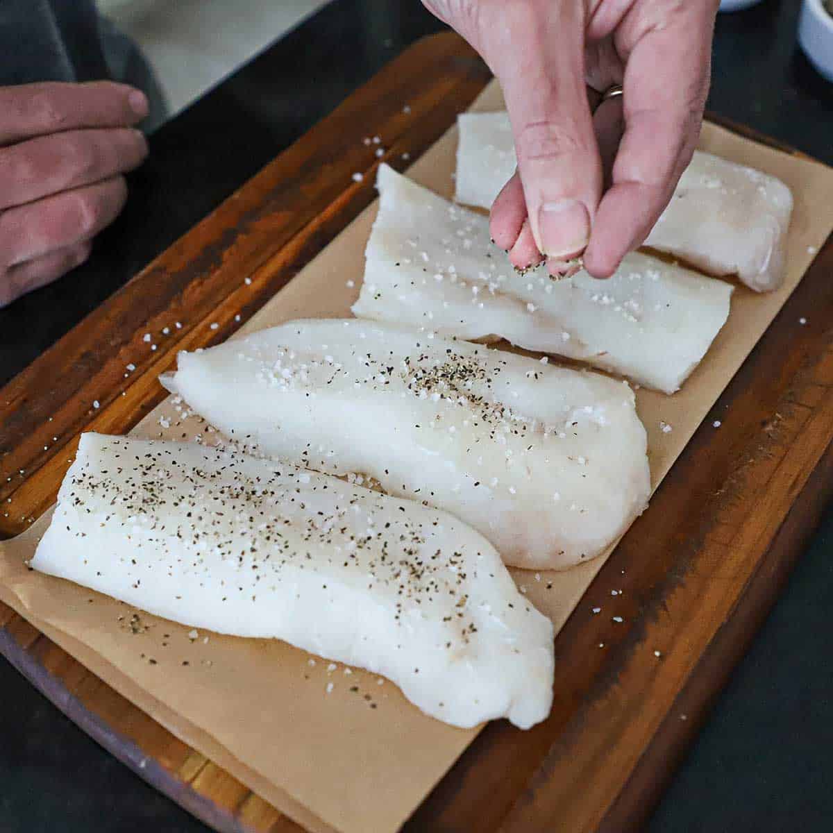A person using his fingers to sprinkle salt and pepper over four uncooked filets of cod resting on brown paper on a small wooden cutting board.