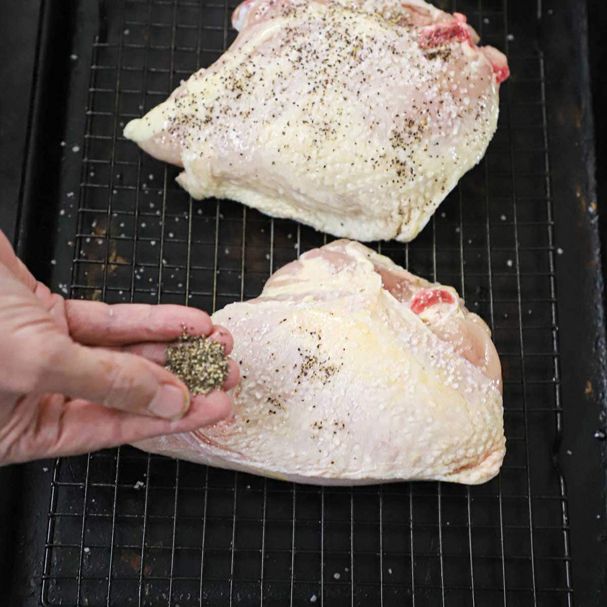 A person using his fingers to sprinkle black pepper over the top of an uncooked chicken breast that is resting on a baking rack in a baking sheet.