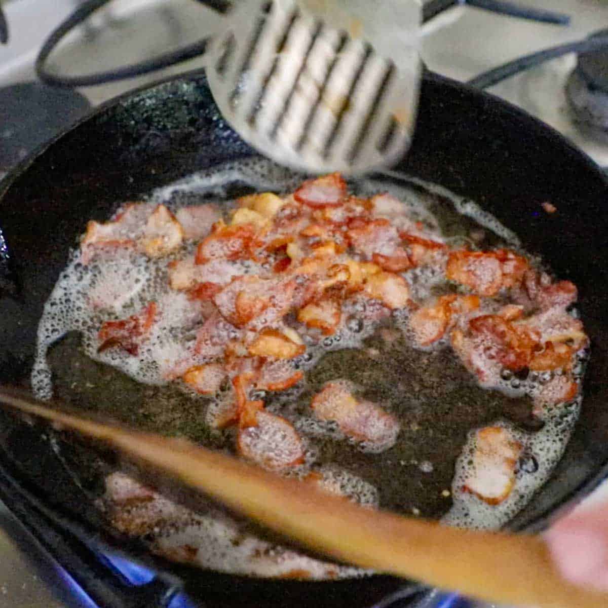 A person using a metal spatula to stir chopped bacon that is being cooked over a gas stove.