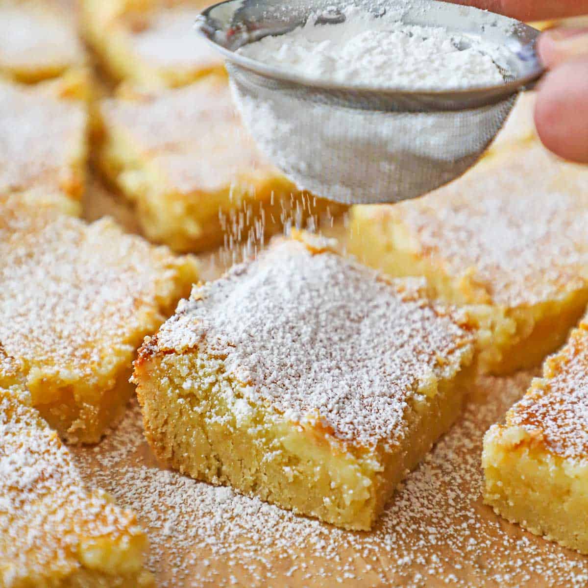 A person using a small sifter to dust powdered sugar over squares of Gooey Butter Cake resting on brown paper.