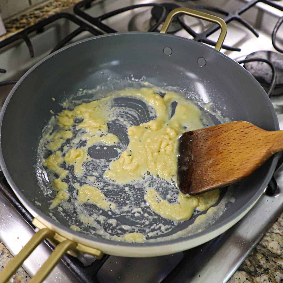 A person using a wooden spatula to stir a roux in a large skillet on a gas stove.