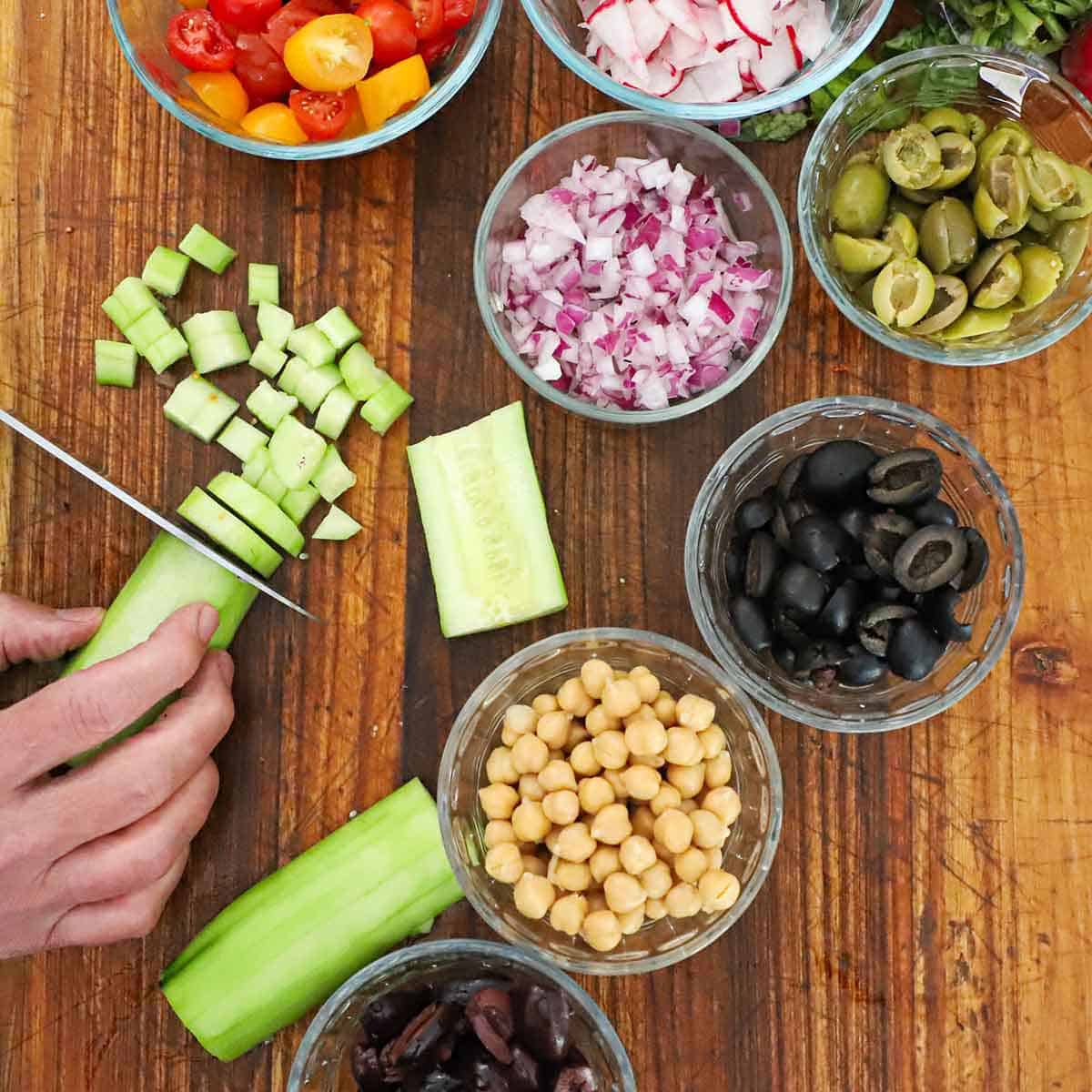 A person slicing a peeled cucumber on a cutting board with small glass bowls filled with various types of olives, garbanzo beans, onions, and tomatoes nearby.