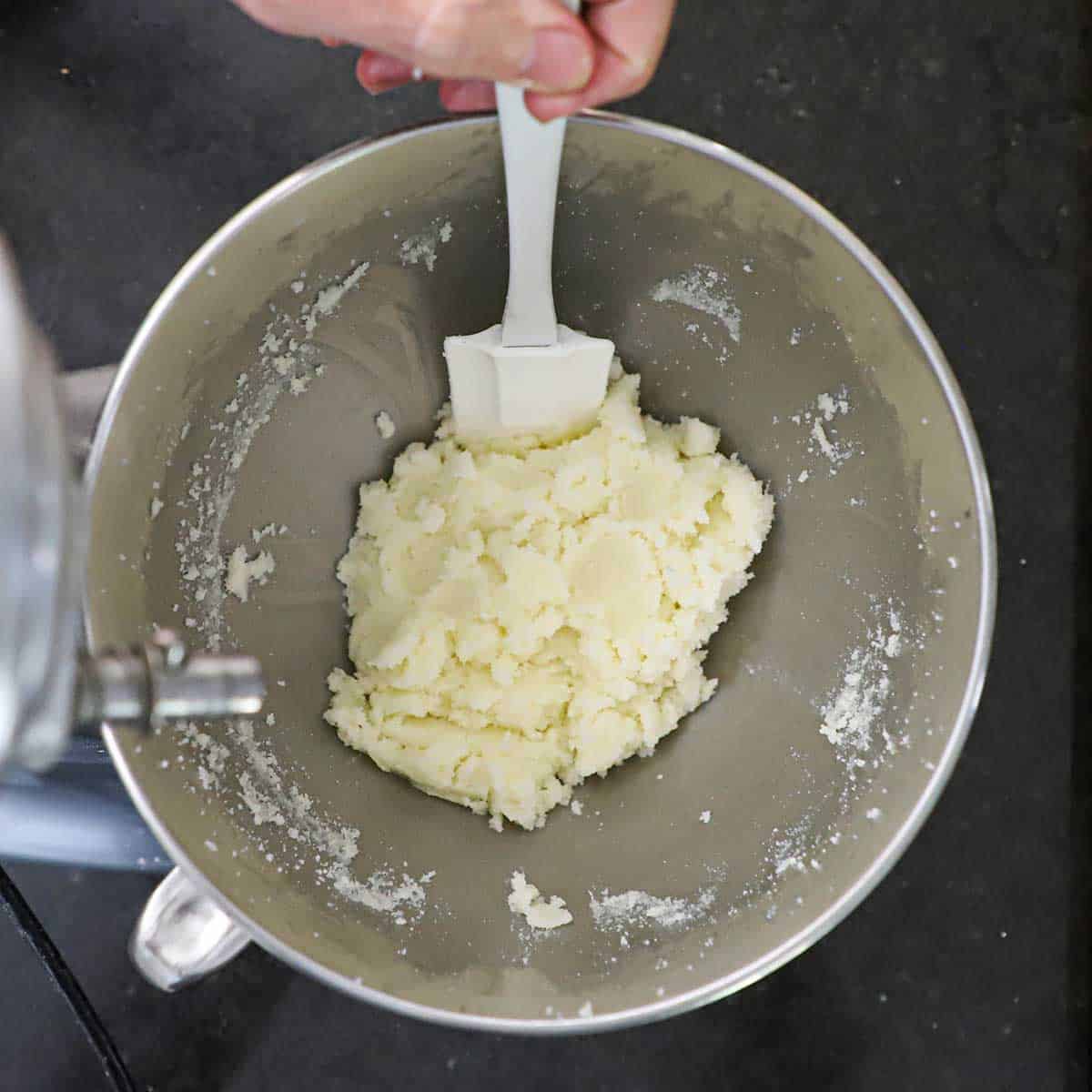 A person using a white spatula to scrape down creamed butter and sugar from the inside of a mixer bowl.