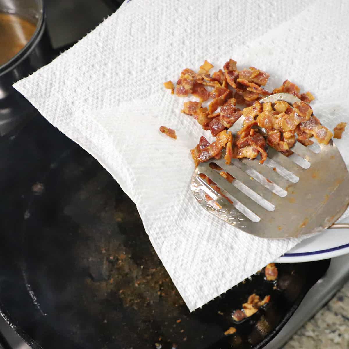 A person using a metal slotted spatula to transfer cooked chopped bacon from a cast-iron skillet to a plate lined with paper towels.