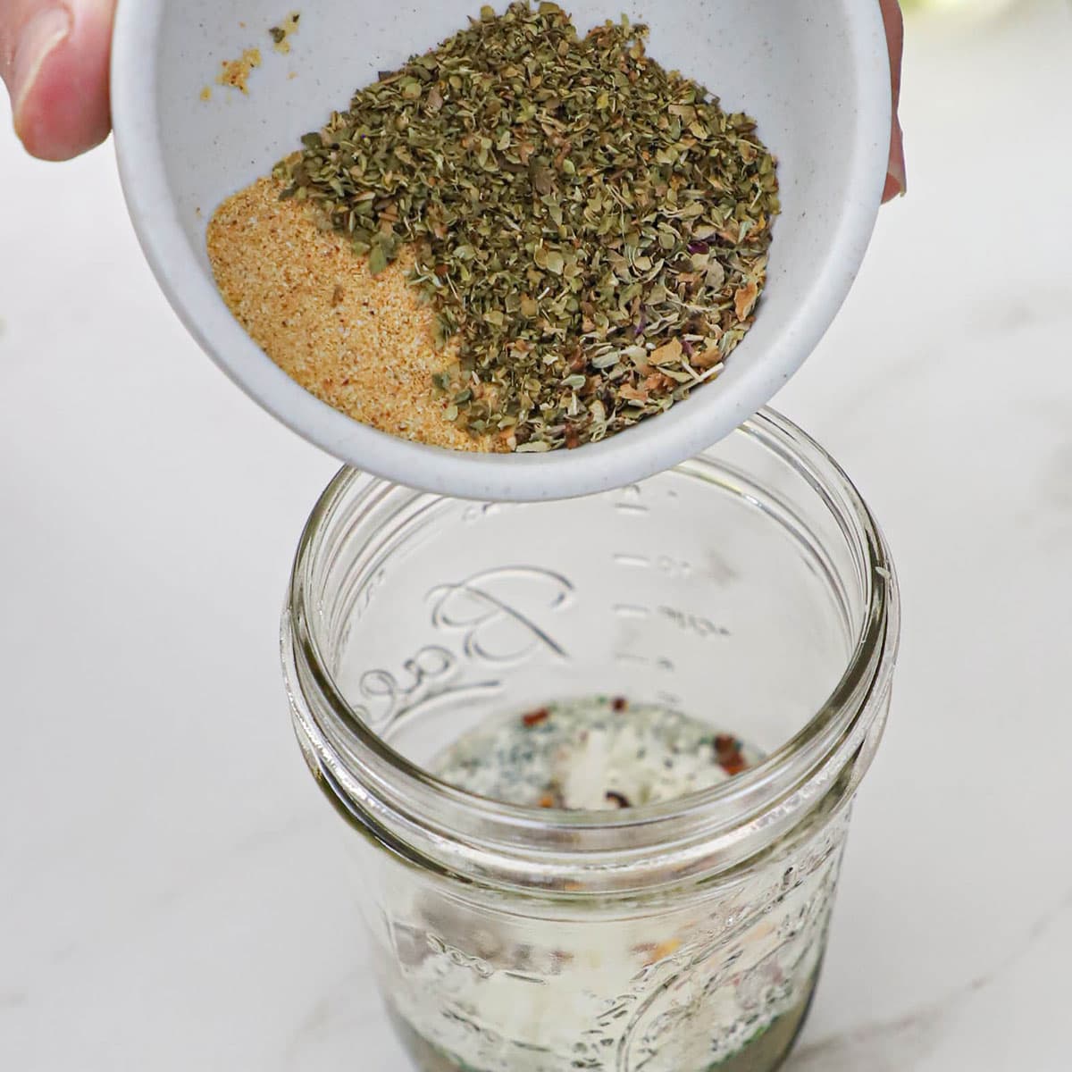 A person dumping dried herbs and onion powder from a small white bowl into a glass jar filled with the ingredients for homemade Italian dressing without the oil added yet.