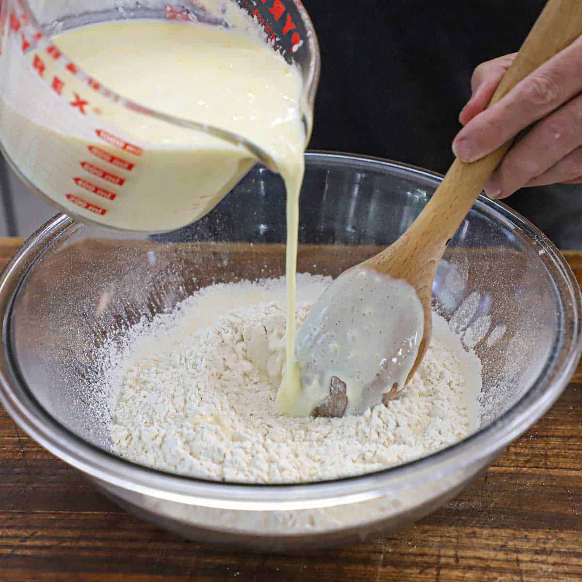 A person pouring buttermilk from a glass measuring cup into a large glass bowl that is filled with dry waffle ingredients.