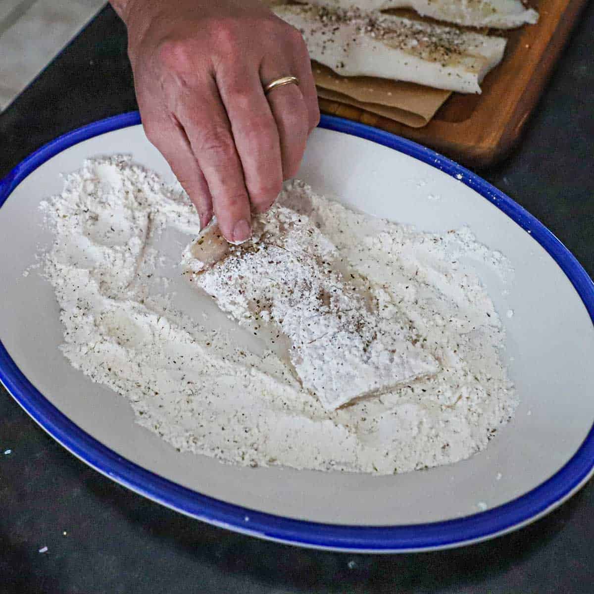 A person dredging a seasoned filet of cod in seasoned flour on a white and blue oval platter.