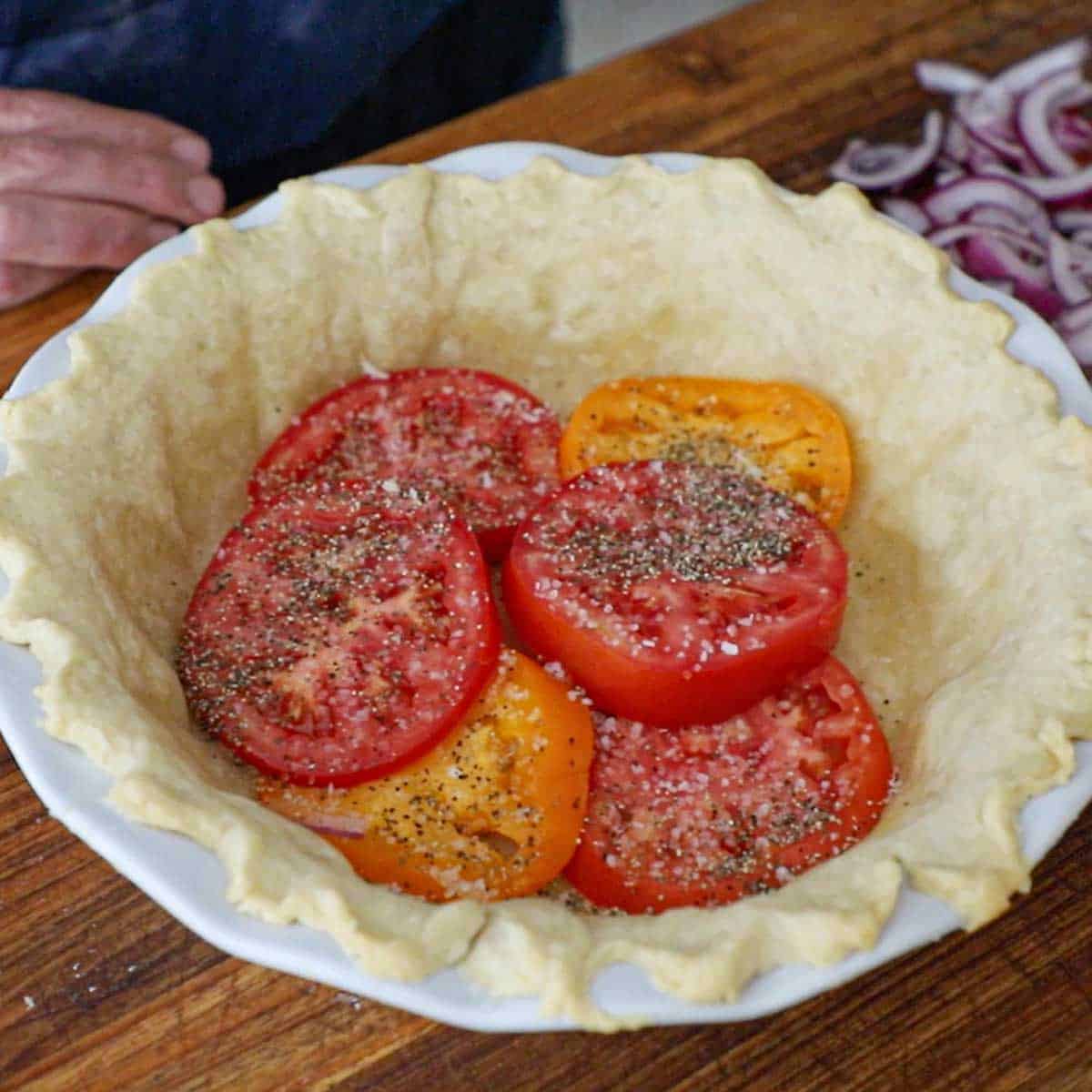 A layer of sliced heirloom tomatoes that have been seasoned with salt and pepper are resting on the bottom of a partially bake pie crust in a pie dish.