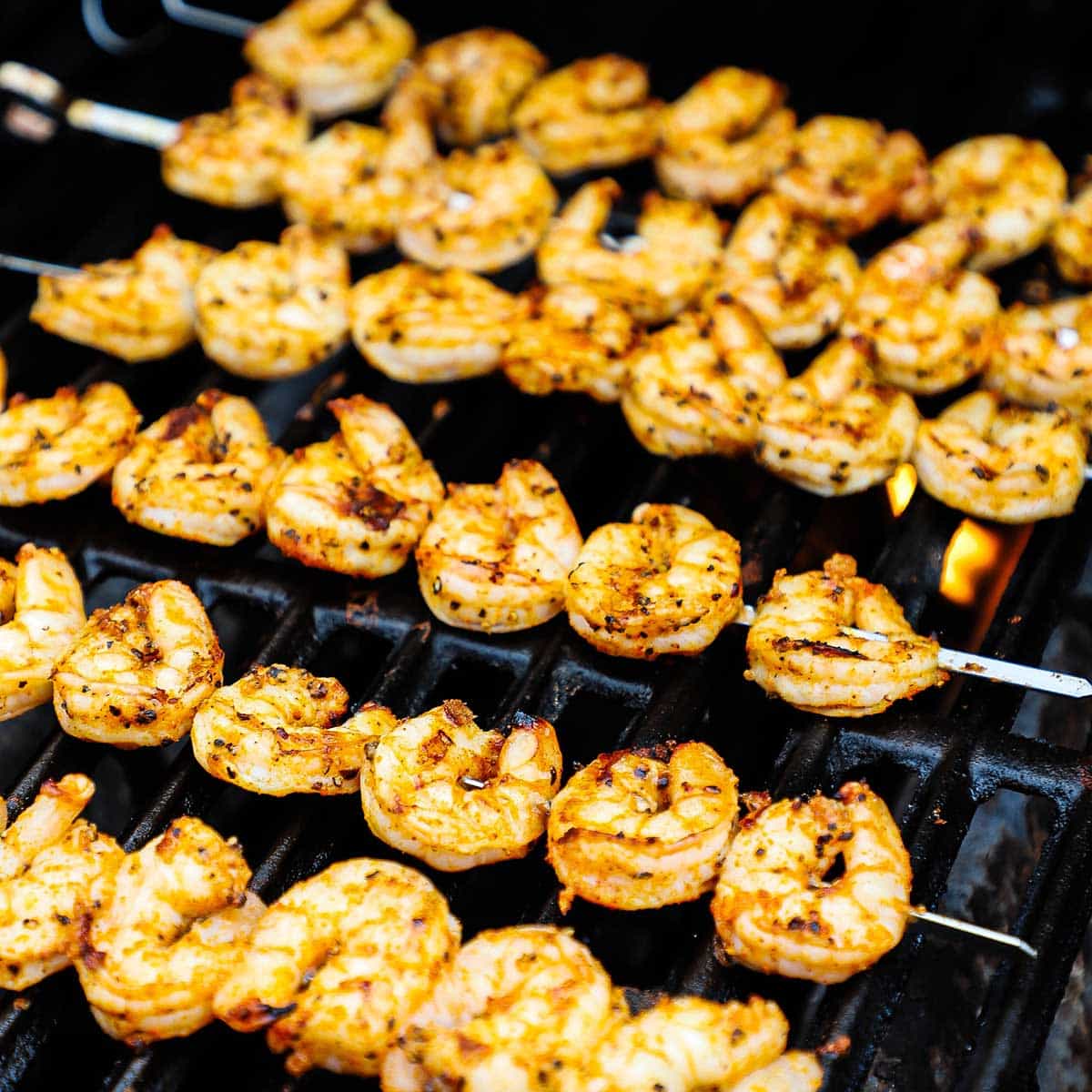 Six rows of seasoned shrimp that are on skewers and are being grilled until slightly charred on a gas grill.