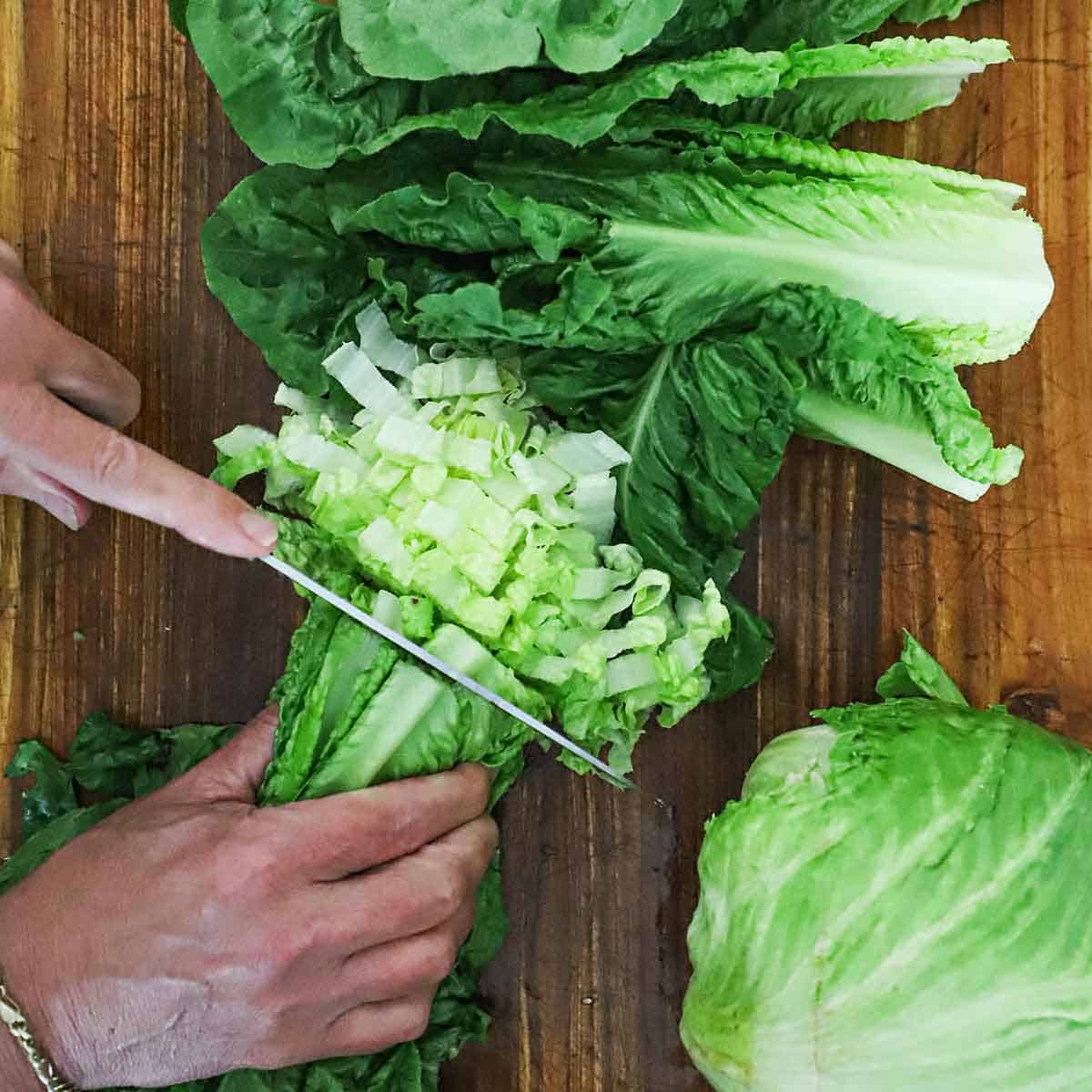 A person using a chef's knife to chop Romaine lettuce on a wooden cutting board.