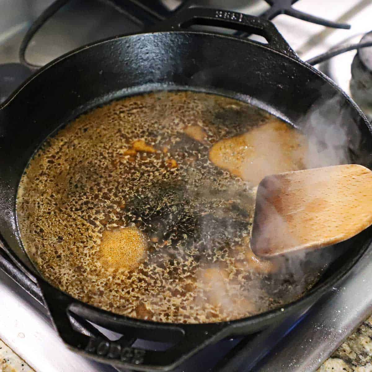 A person using a wooden spatula to stir boiling beef stock in a black cast-iron skillet on a gas stove.
