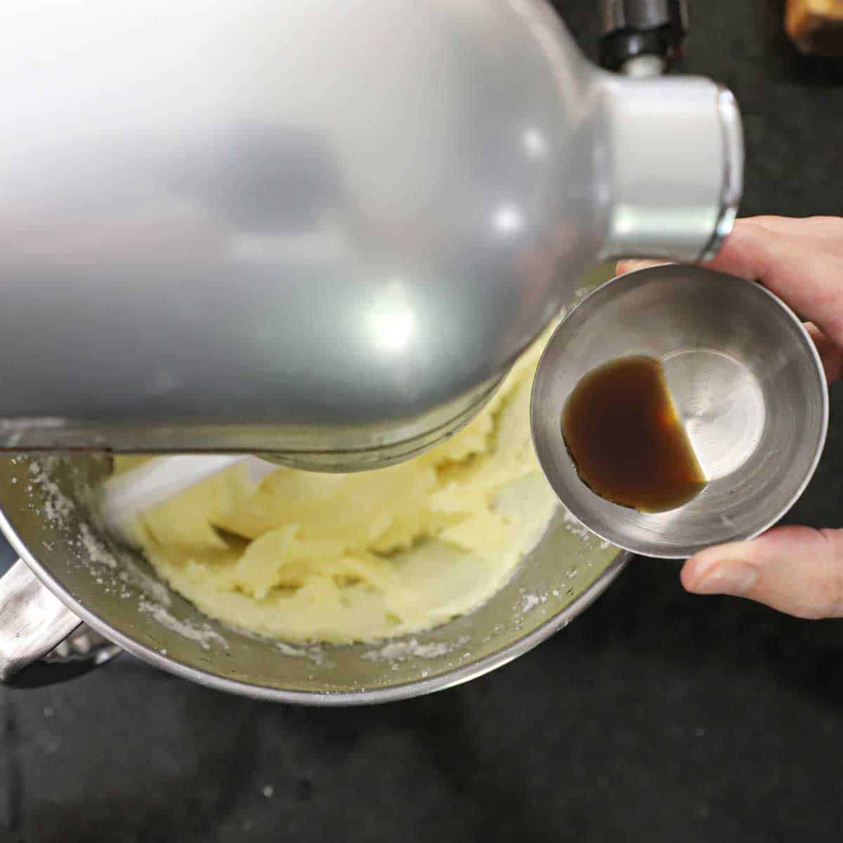 A person holding a small silver bowl filled with a teaspoon of vanilla extract over the bowl of a stand mixer that is filled with creamed butter and sugar.